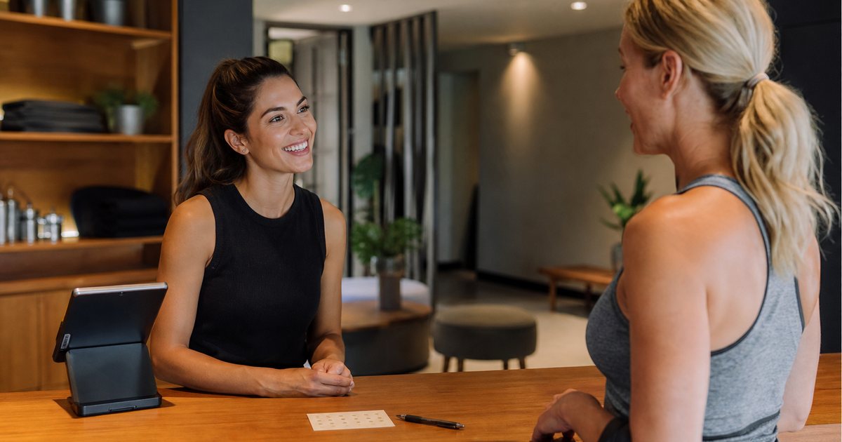 A studio front desk with a card reader, a paper punch card and a pen on the wooden counter, a staff member and a member in workout clothes talking, warm interior lighting