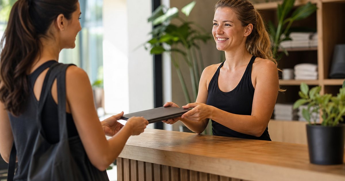 A friendly front desk staff member handing a welcome folder to a new member in a bright studio lobby with green plants and a wooden counter, both smiling, morning light