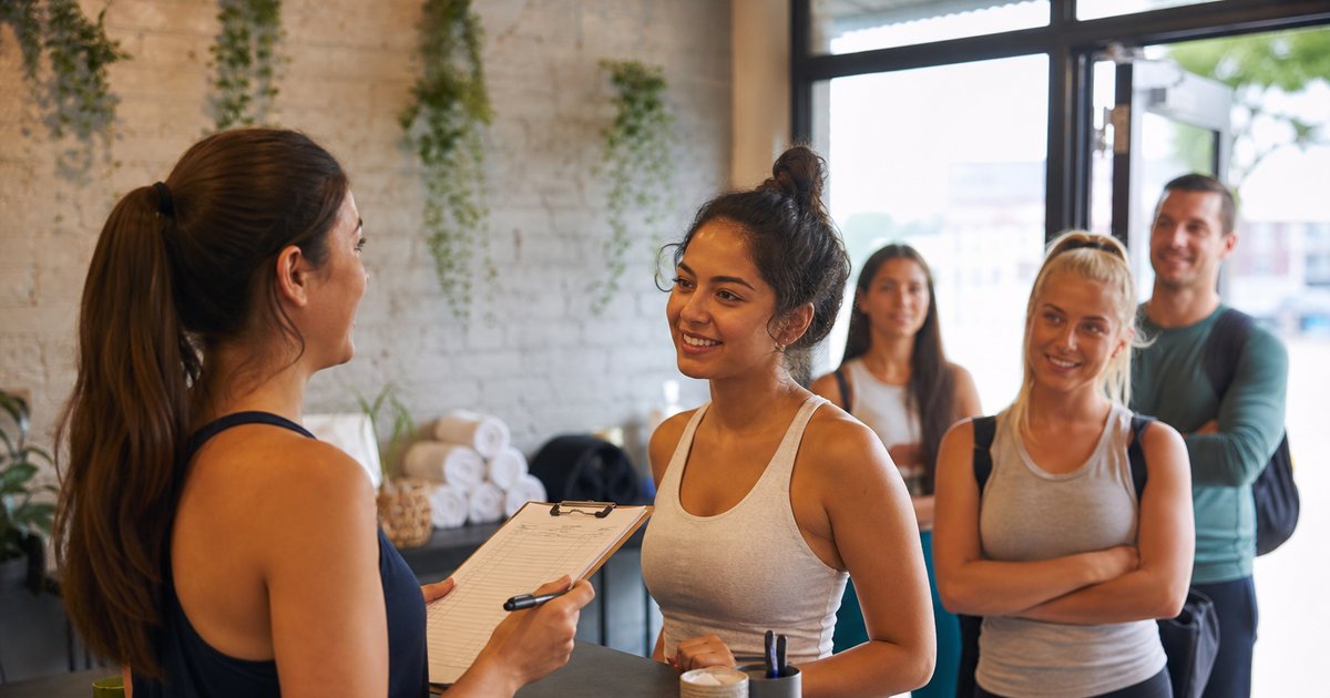A bright boutique fitness studio front desk in the morning, a staff member holding a paper class roster on a clipboard while several members in athletic wear wait near the door